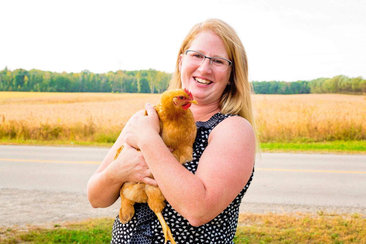 Smiling blonde woman holding a golden chicken in a sunny farm field setting.