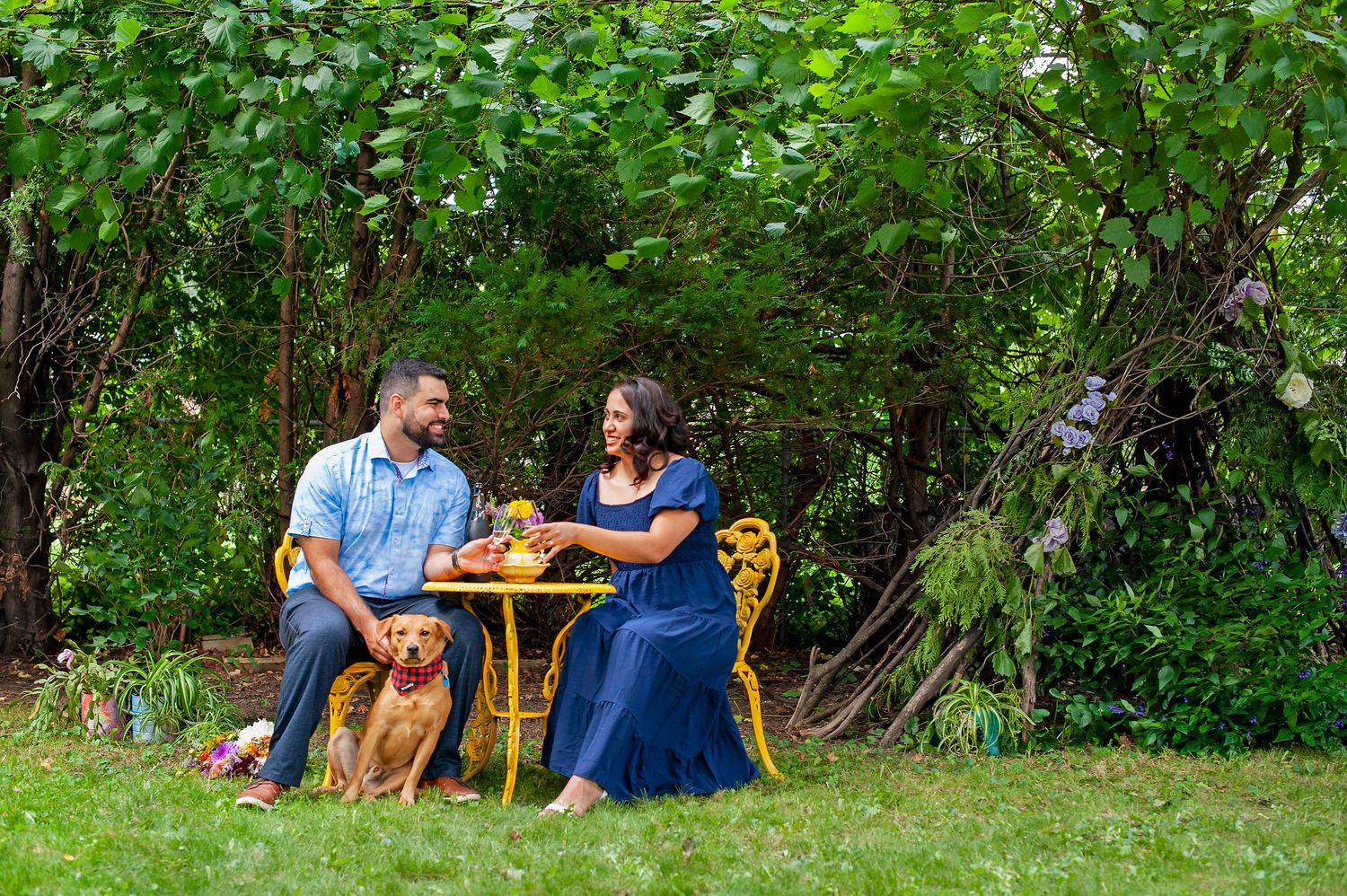 Couple sharing a romantic moment at a yellow table with their dog in a garden.