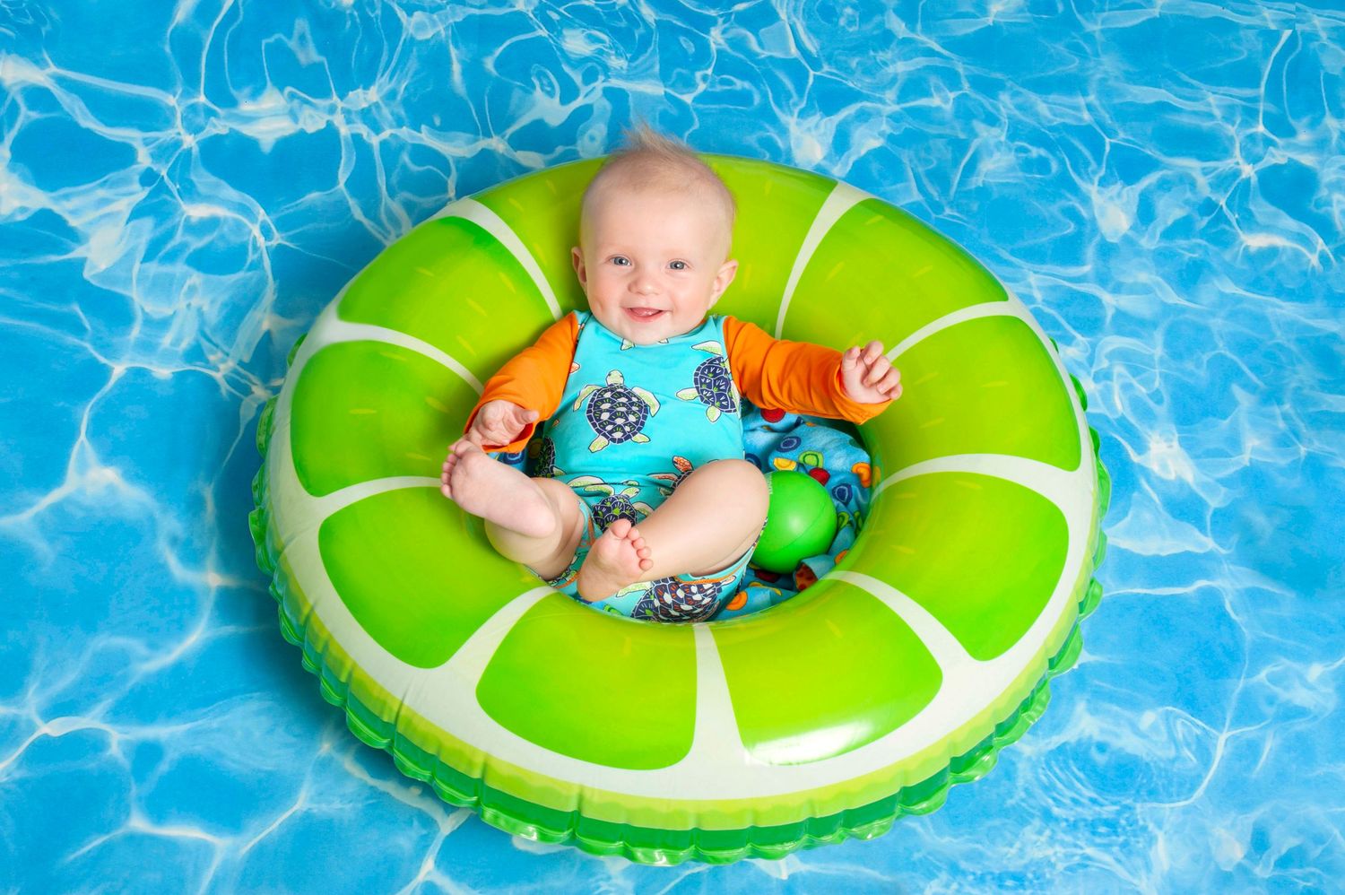 Smiling baby sitting in a green lime inflatable ring float in a bright blue swimming pool.