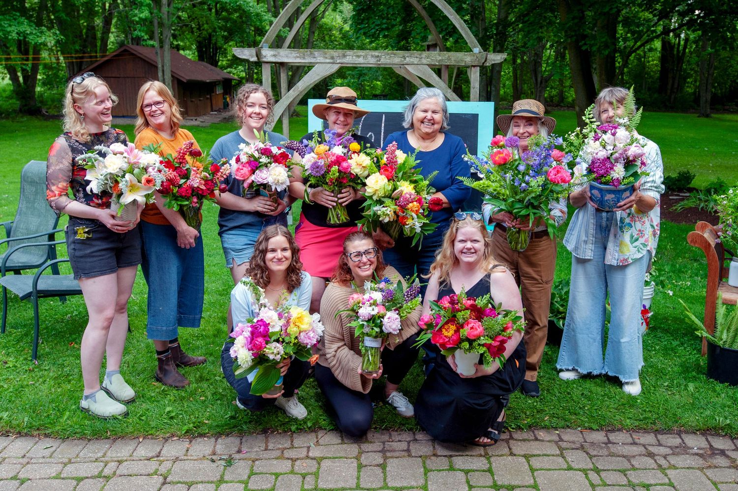 Group of women posing outdoors holding colorful flower bouquets in a lush green garden setting.