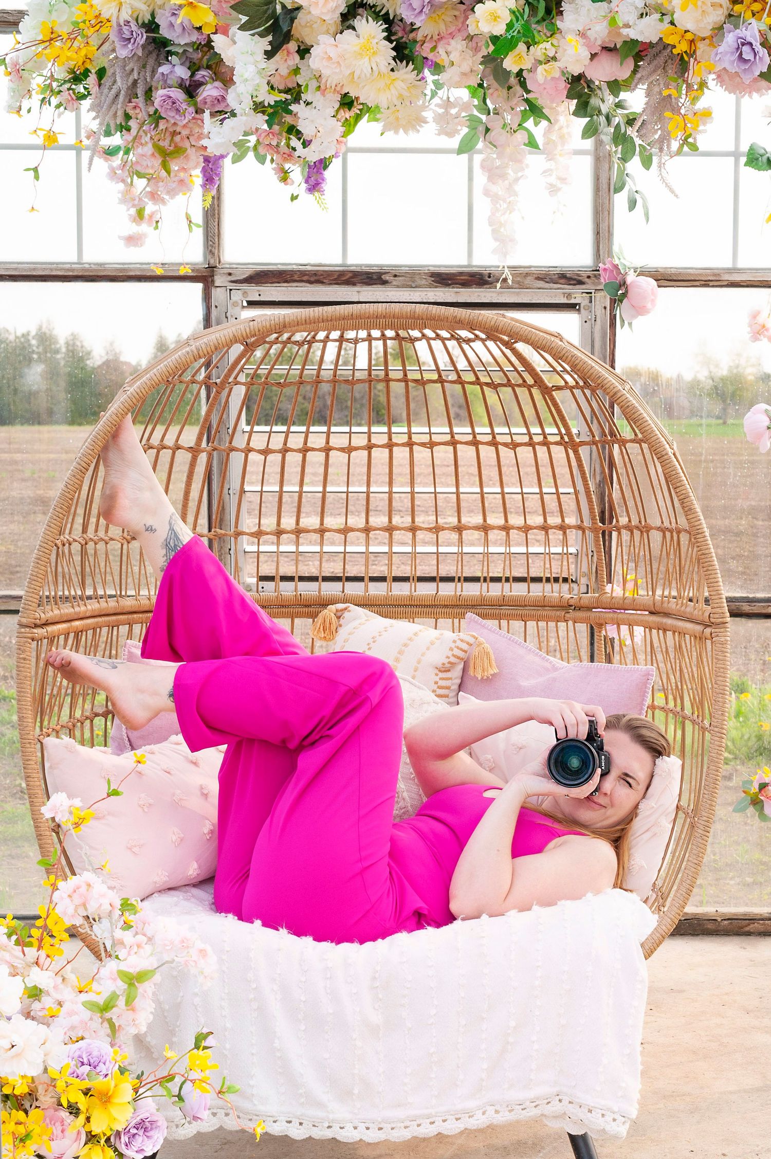 Woman in pink jumpsuit lying in wicker egg chair holding camera among spring flowers in bright greenhouse setting.