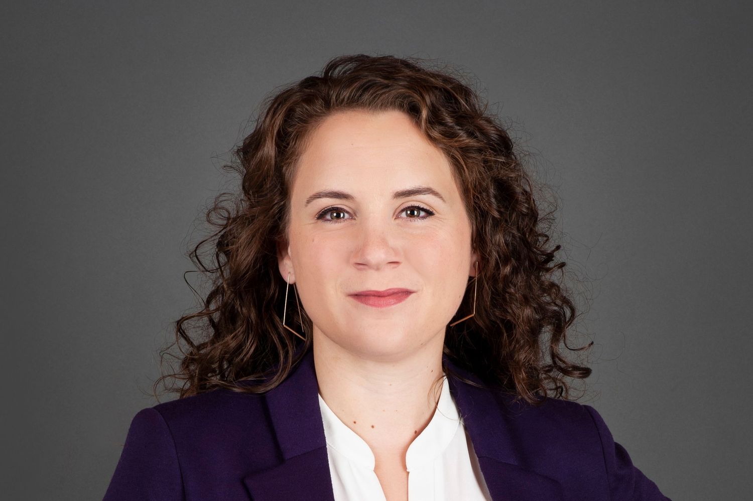 Professional woman with curly hair in purple blazer against gray studio background.