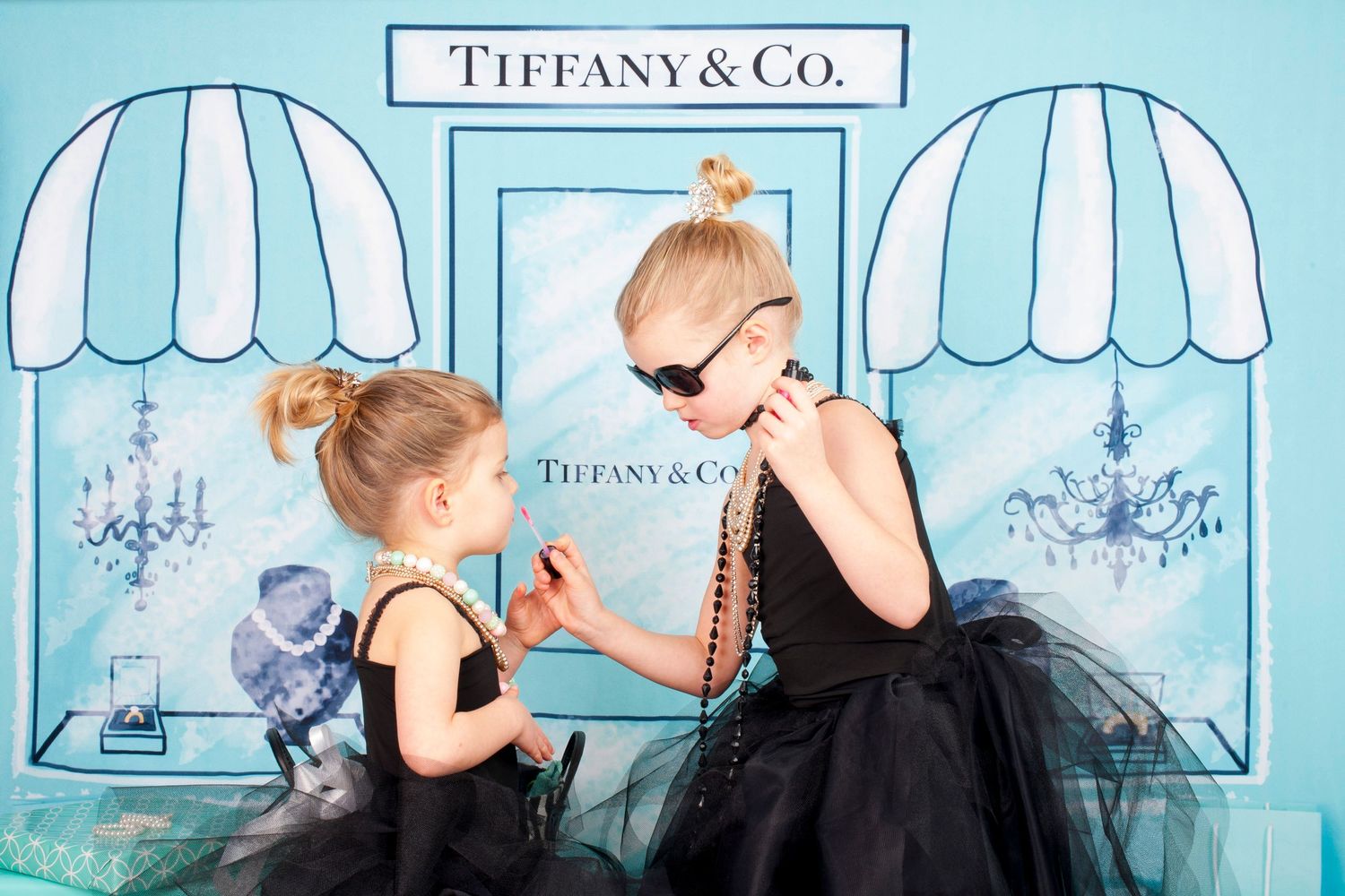 Two little girls in black tutus playing dress-up in front of a painted Tiffany and Co storefront backdrop.