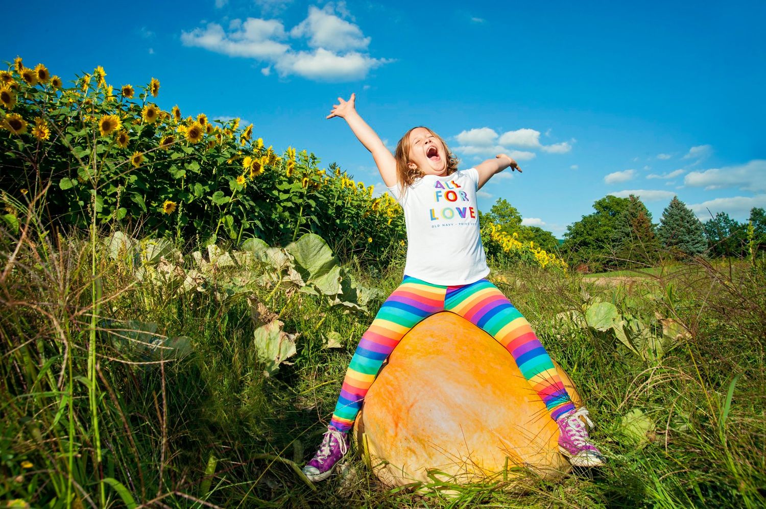 Girl in rainbow leggings sits on giant pumpkin near sunflowers, arms raised joyfully under blue sky.