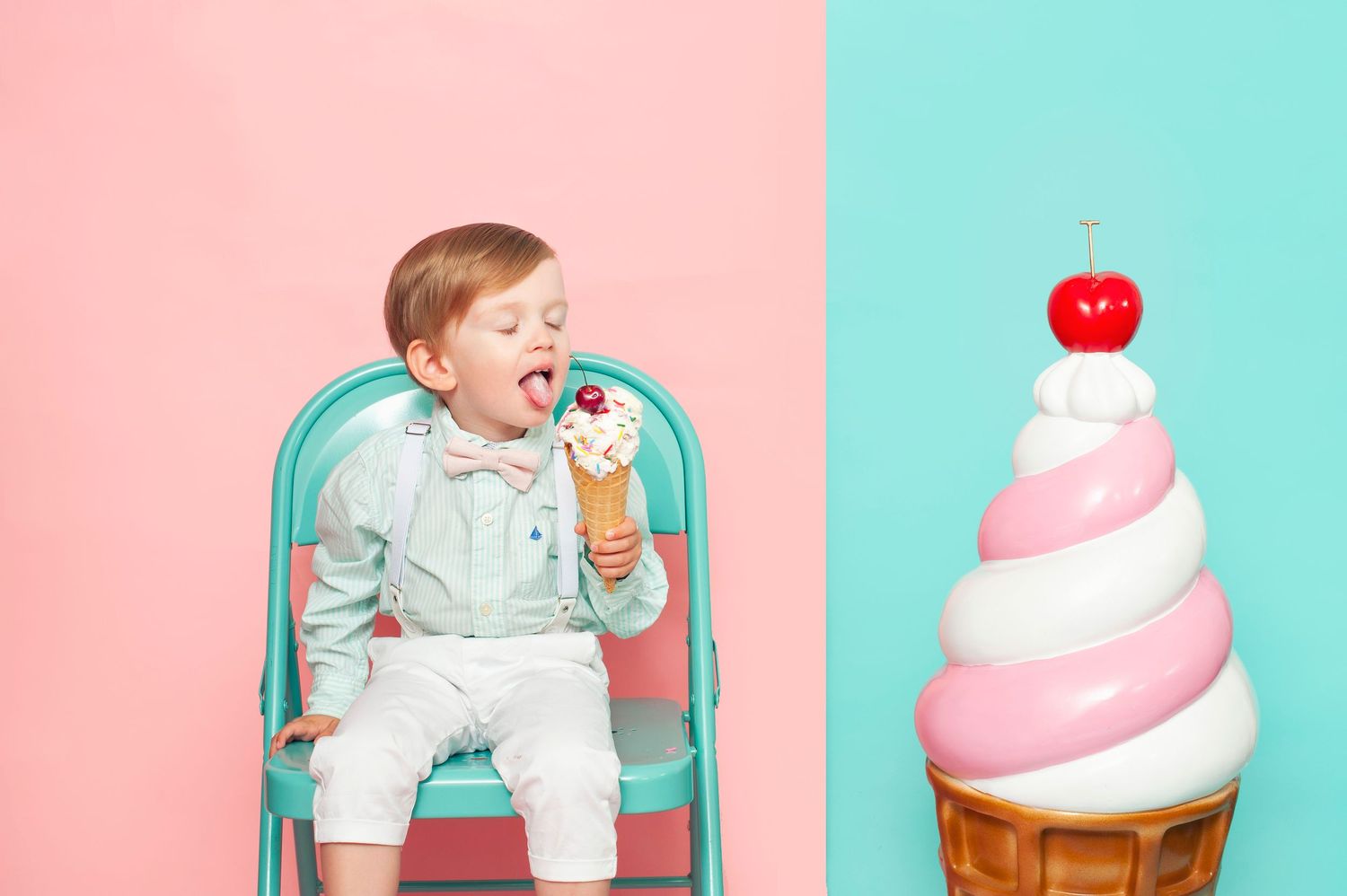 Toddler boy licking ice cream cone while sitting on teal chair beside large decorative ice cream on pink blue backdrop.
