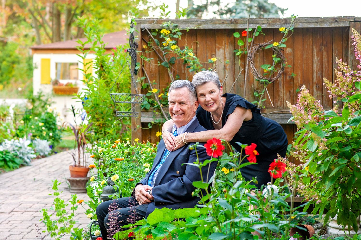 Elderly couple smiling in a lush garden surrounded by colorful flowers and greenery.