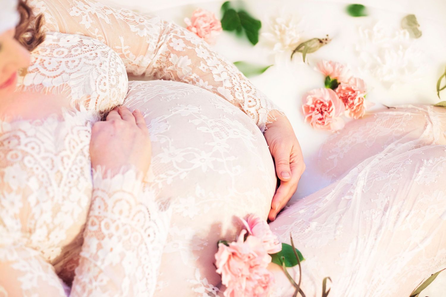 Close-up of pregnant woman in white lace dress cradling baby bump surrounded by pink flowers.