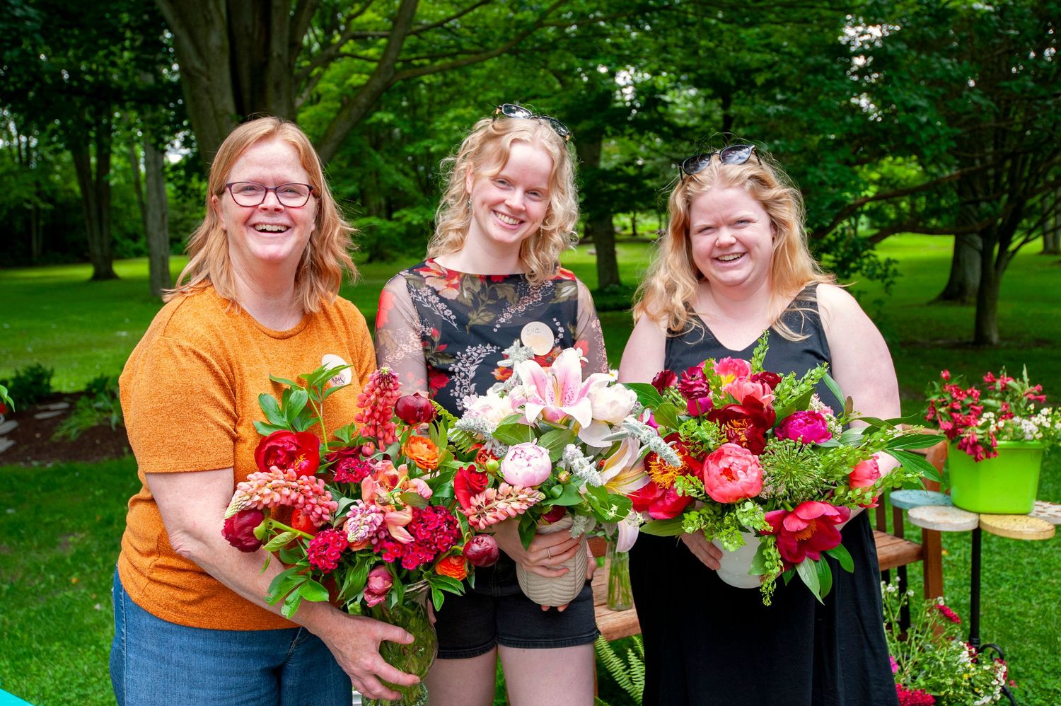 Three women smiling outdoors holding large colorful floral bouquets in a lush green garden setting.