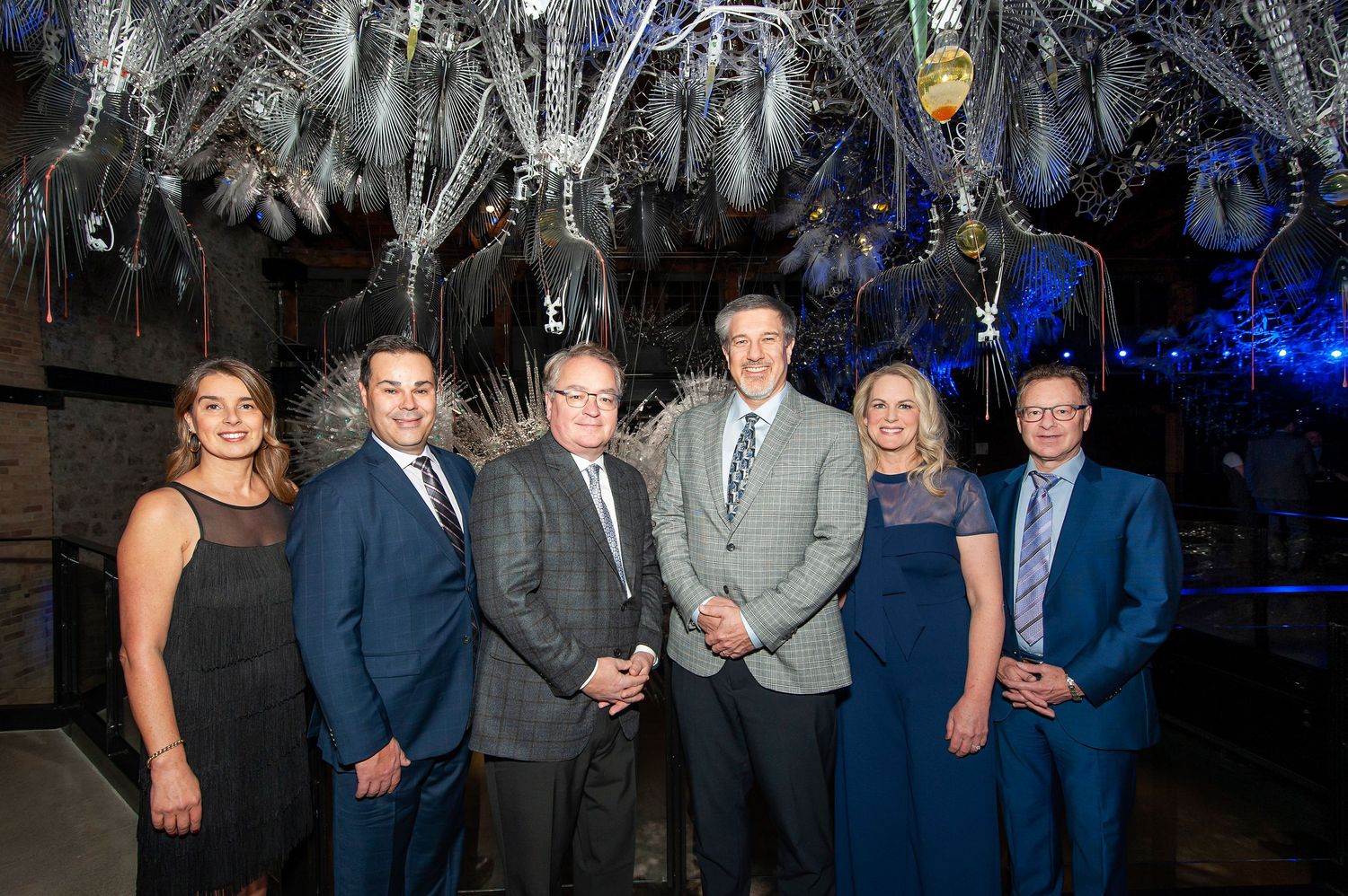 Six professionals pose together at a formal gala event under decorative hanging ornaments.