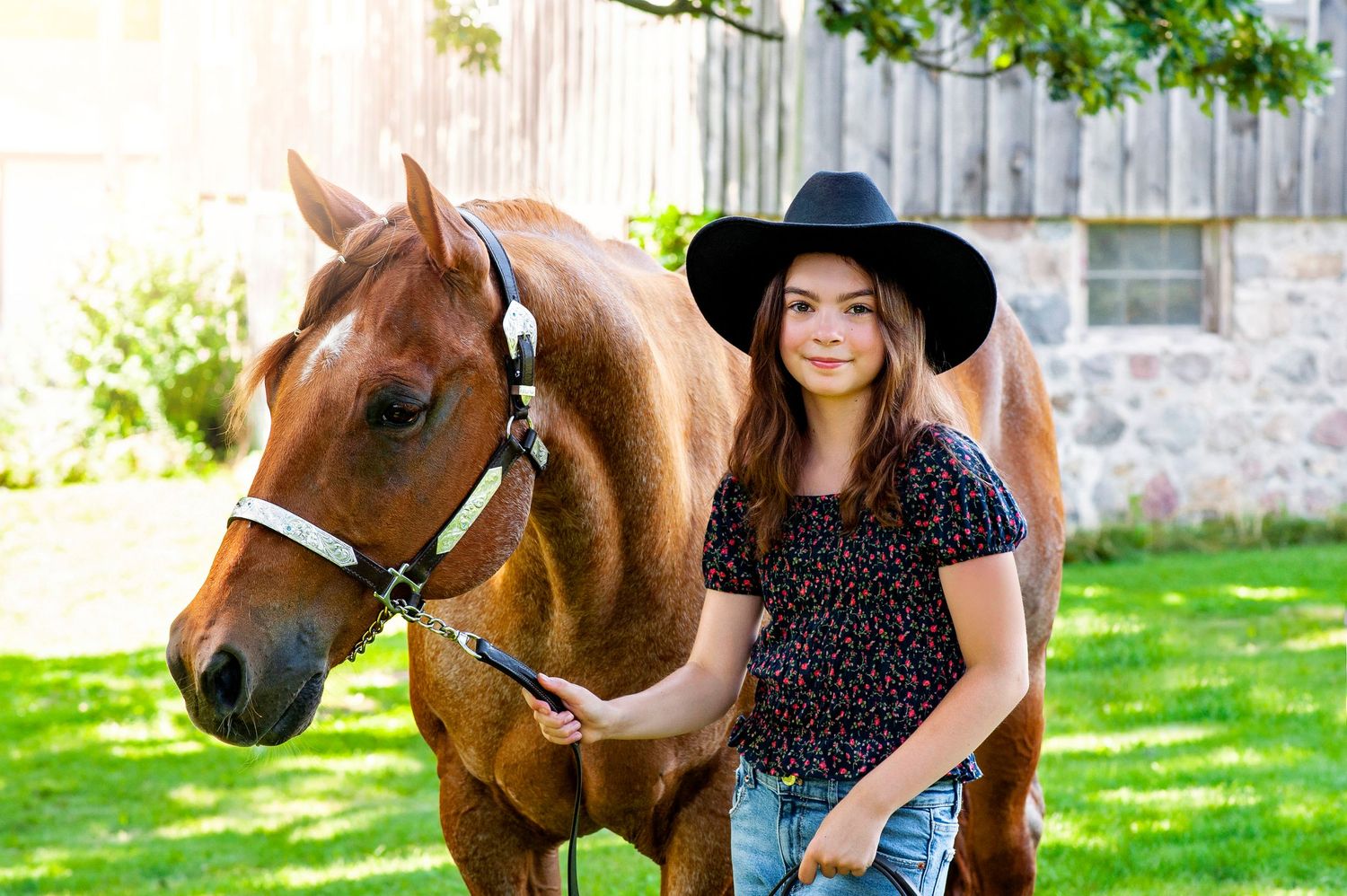 Teen girl in black hat posing with chestnut horse near rustic stone barn outdoors.