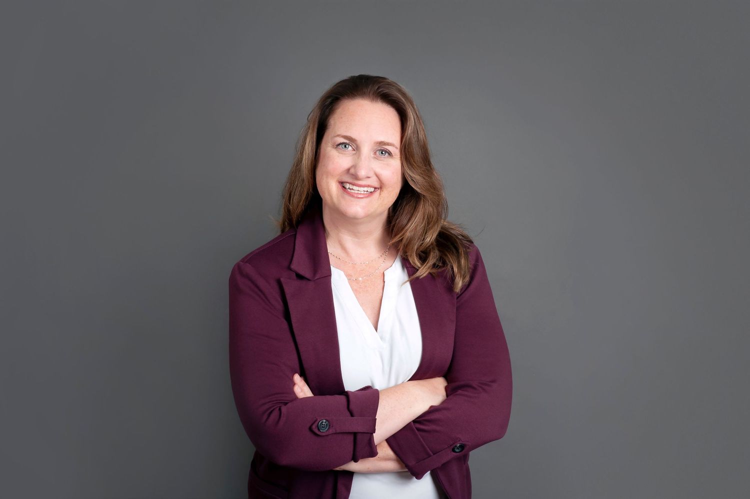 Professional woman in burgundy blazer smiles with arms crossed against gray background.