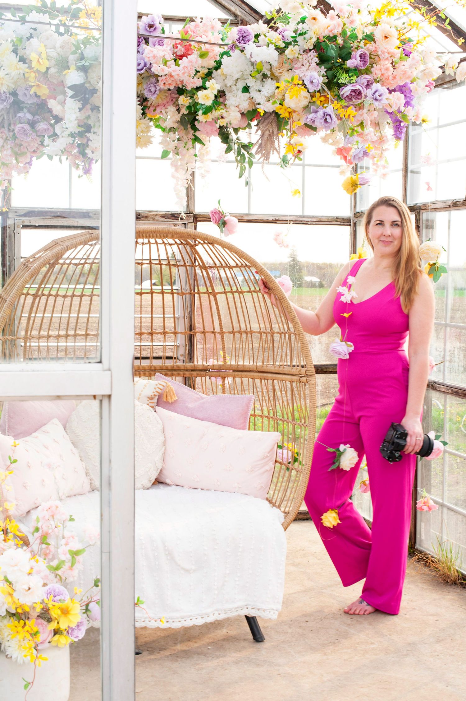 Woman in bright pink jumpsuit holding camera stands beside wicker egg chair in flower-filled greenhouse studio.