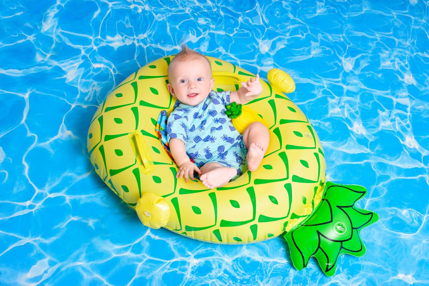 Happy baby relaxing on a pineapple inflatable float in a sparkling blue swimming pool.