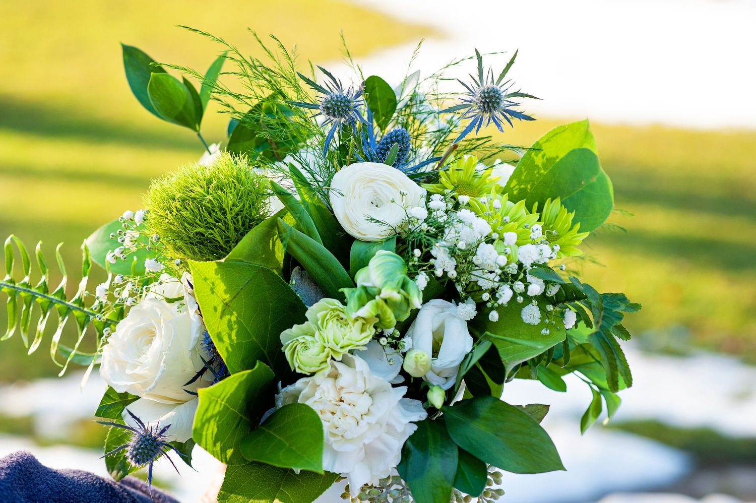 Elegant bouquet of white ranunculus, green dianthus, blue eryngium, and mixed foliage held outdoors.