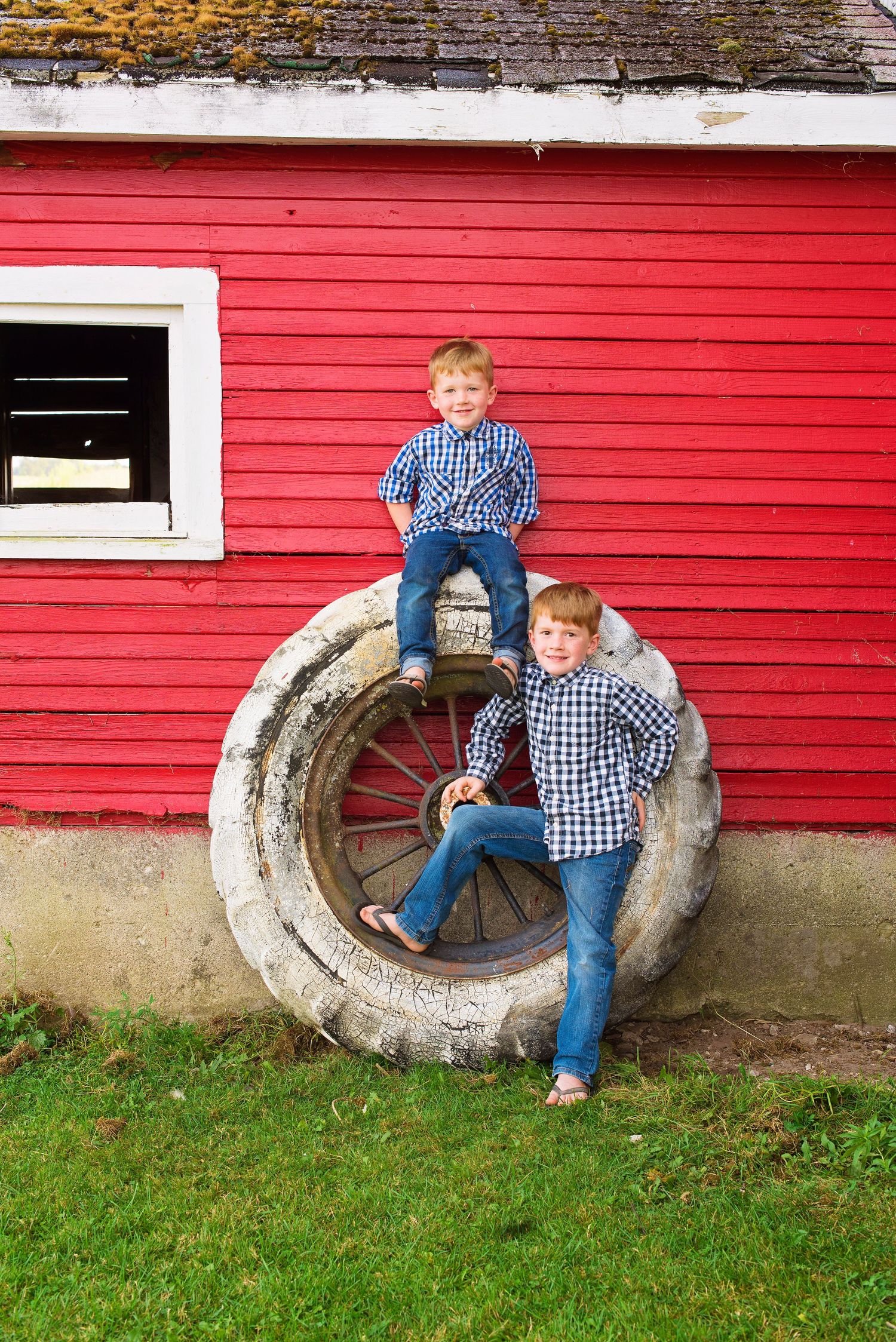 Two young boys in plaid shirts posing on a large vintage tire against a red barn wall.