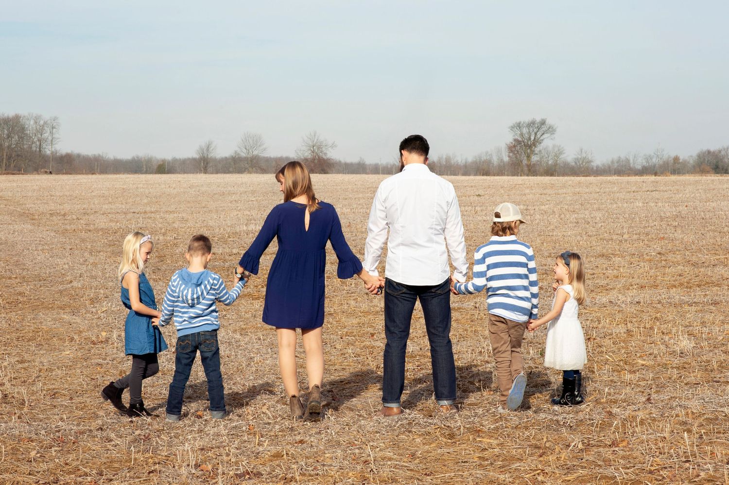 Family of six holding hands walking through a harvested field on a clear autumn day, viewed from behind.