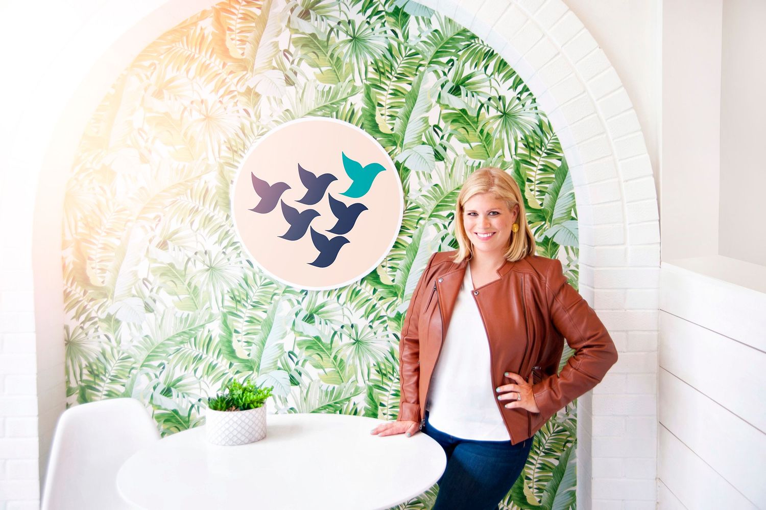 Smiling blonde woman in a brown leather jacket posing beside a tropical leaf wallpaper with bird artwork.