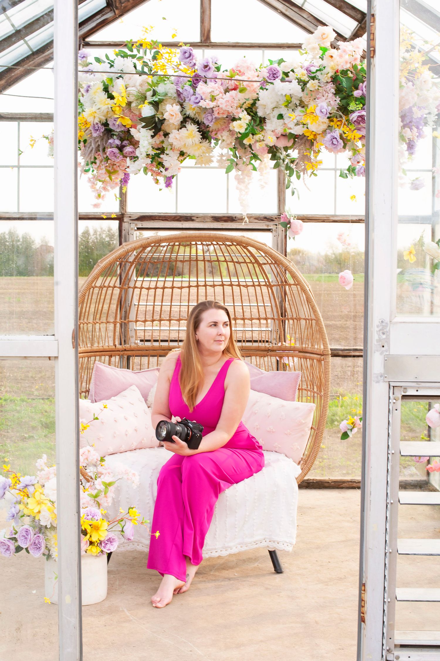 Woman in pink jumpsuit holding camera sits in wicker egg chair surrounded by colorful floral arrangements in greenhouse.