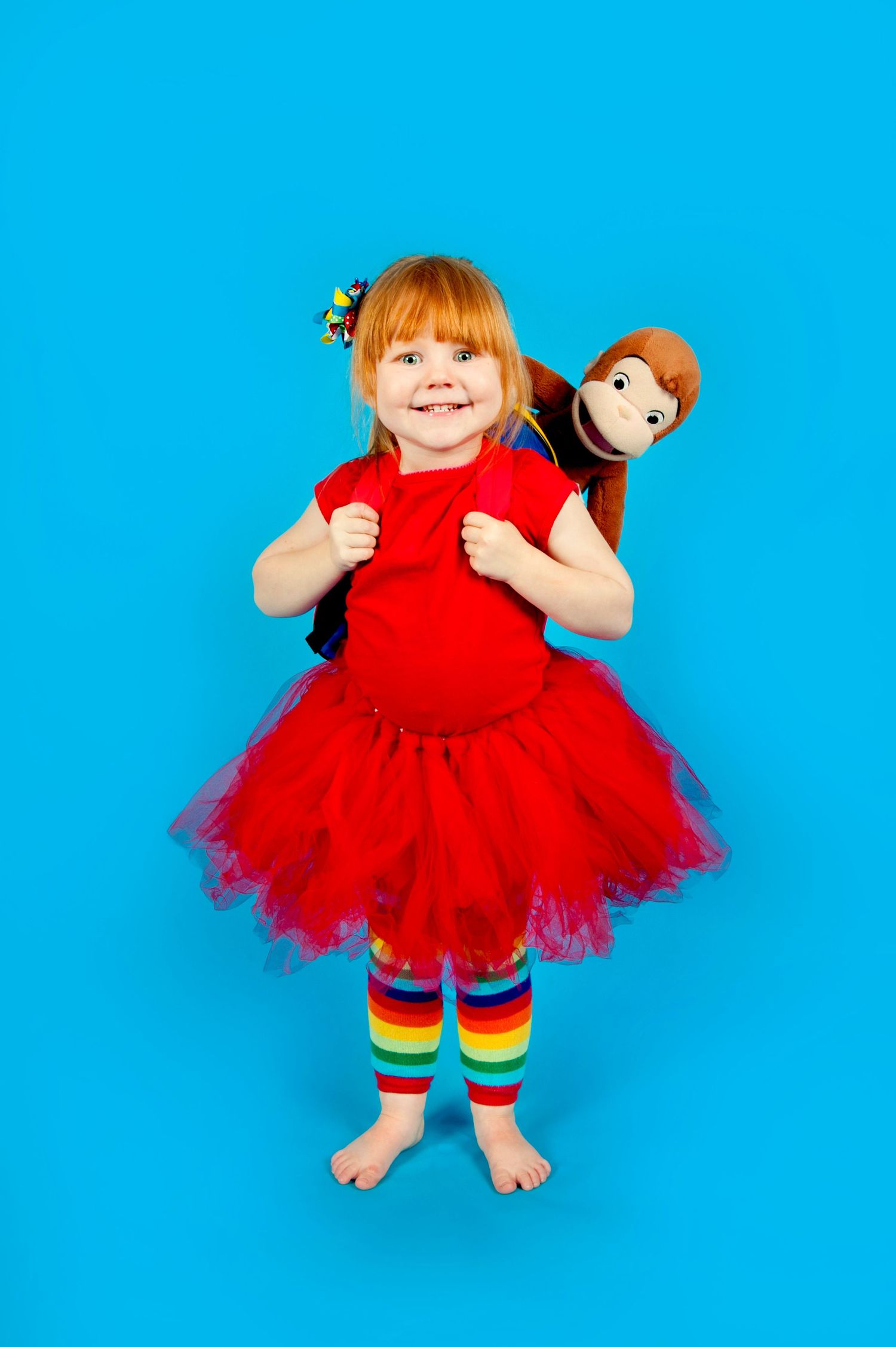 Smiling redhead girl in red tutu dress and rainbow leggings holding a monkey stuffed animal backpack on blue background.