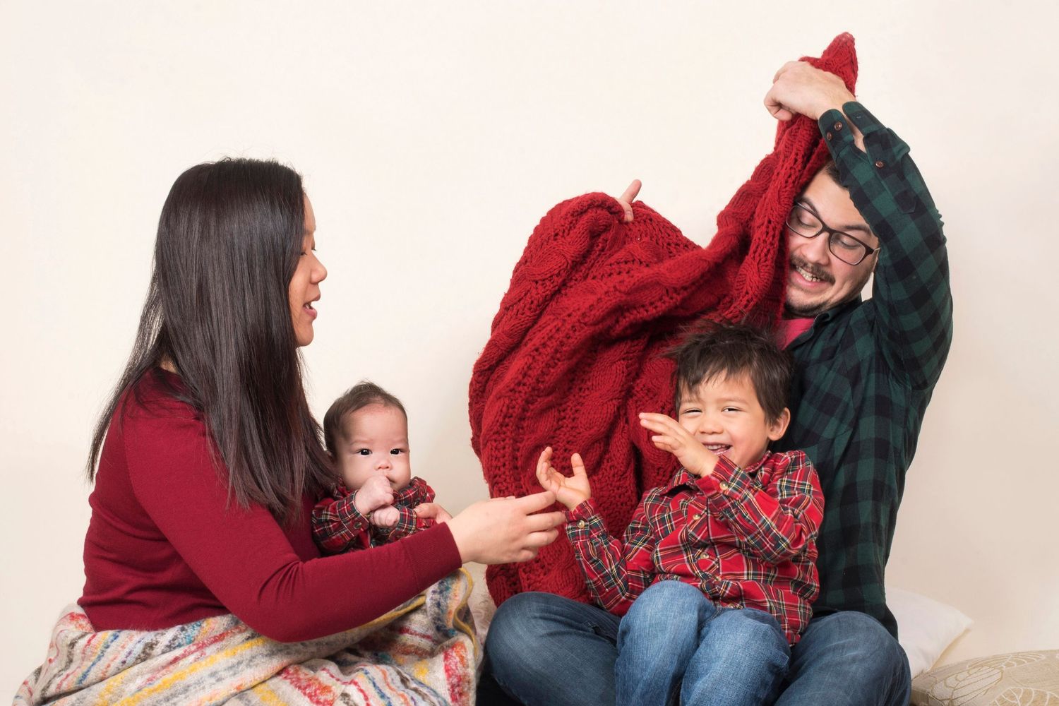 Happy family of four playfully sharing a red knit blanket during a photo session.