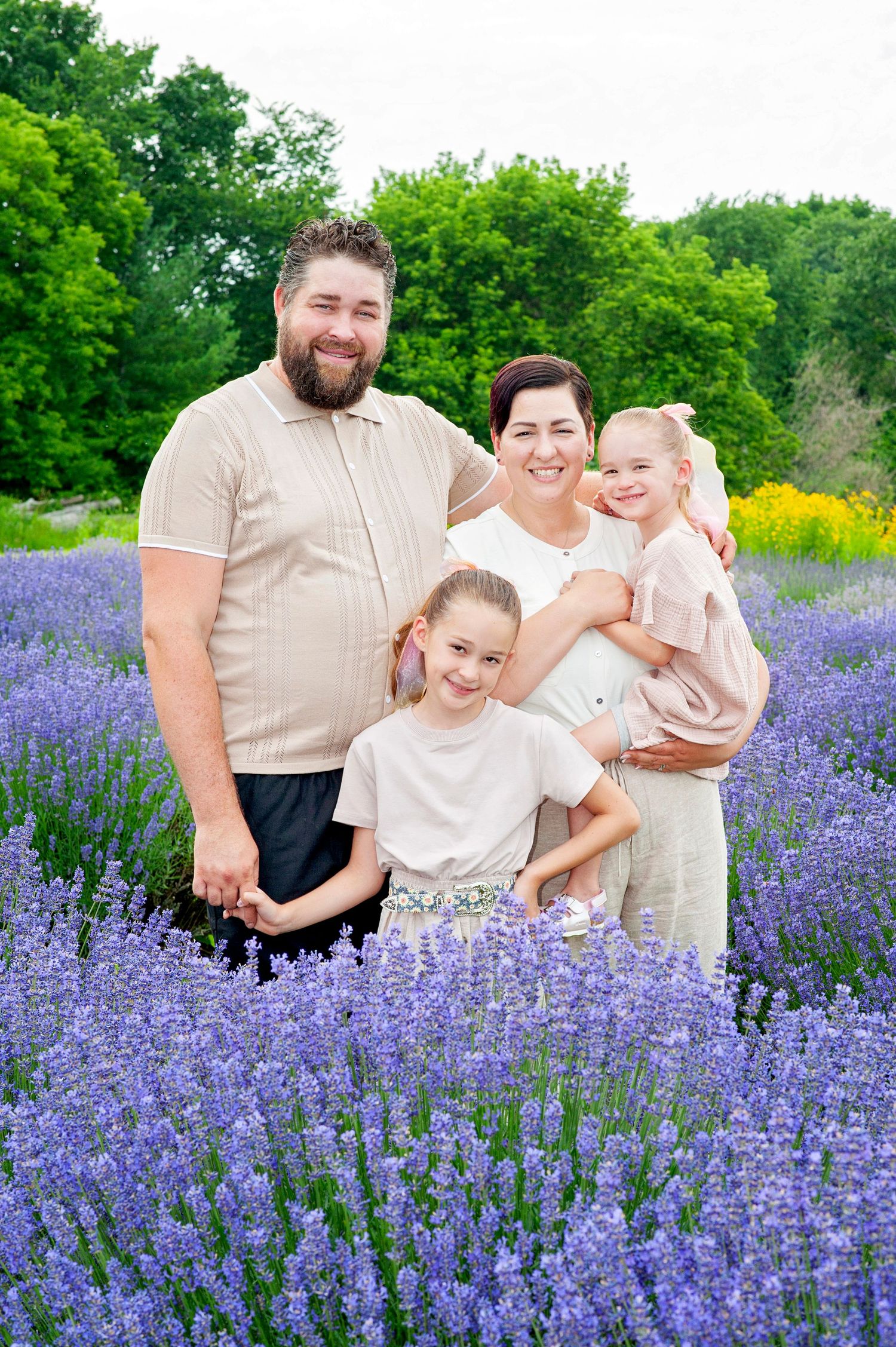 Happy family of four poses together in a vibrant purple lavender field with green trees behind them.