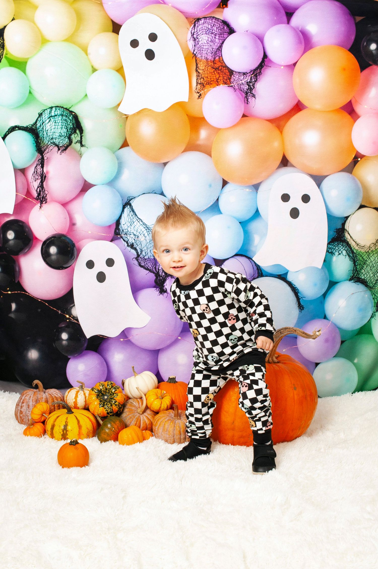 Playful toddler in checkered outfit posing with pumpkins and colorful ghost balloon wall.