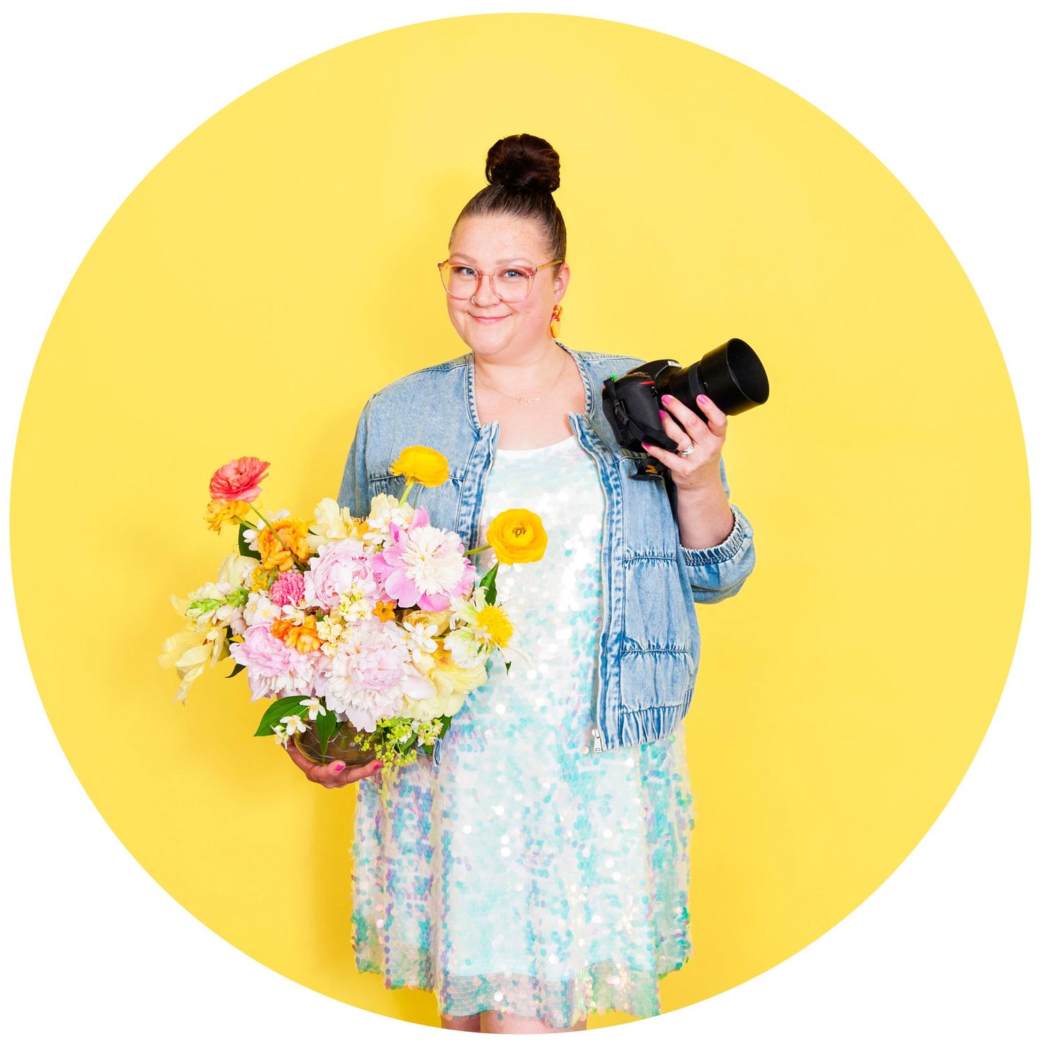 Woman in floral dress holding colorful bouquet and camera, smiling against bright yellow background.