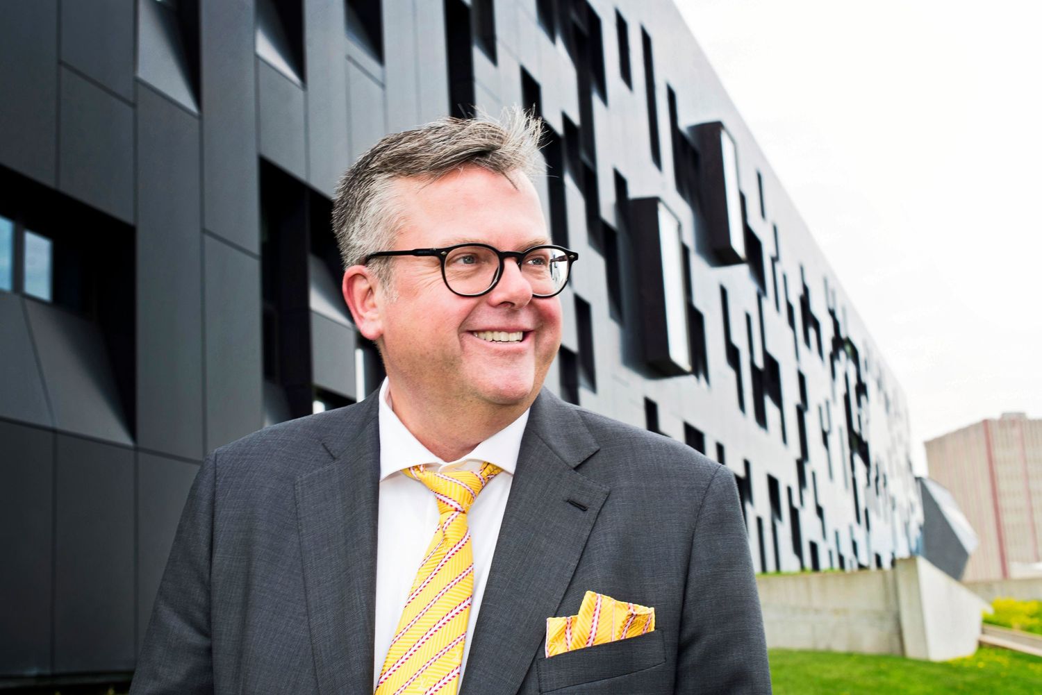 Smiling businessman in gray suit and yellow tie stands outside modern geometric black building exterior.