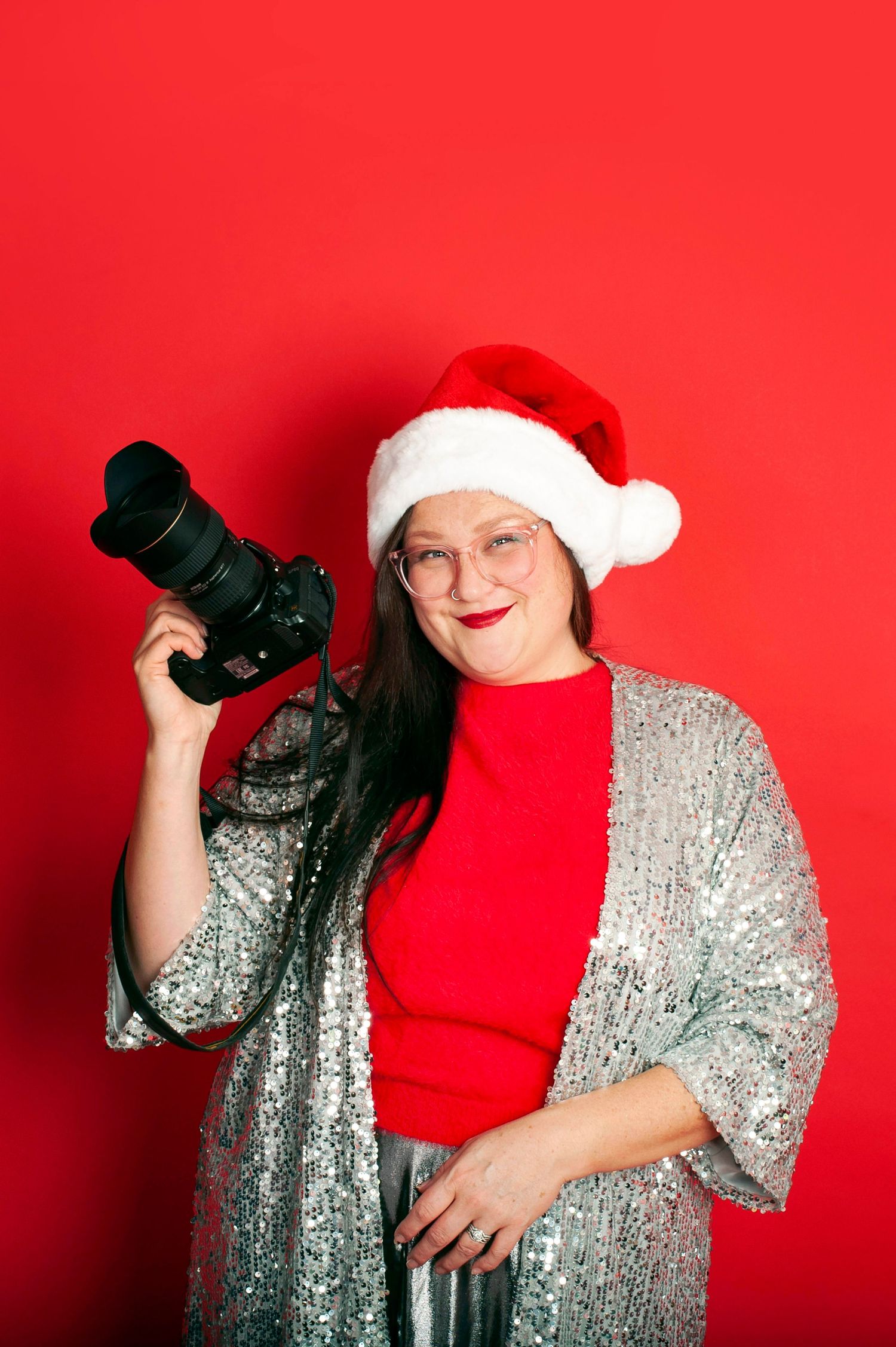 Woman in Santa hat and sequin jacket holds DSLR camera, smiling against red background.