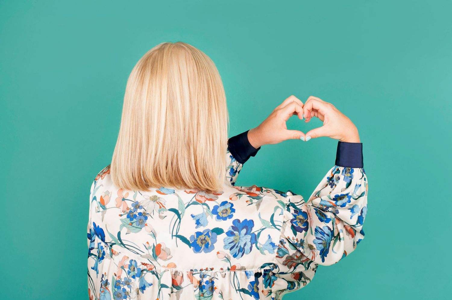 Blonde woman making heart shape with hands wearing floral jacket against teal background.