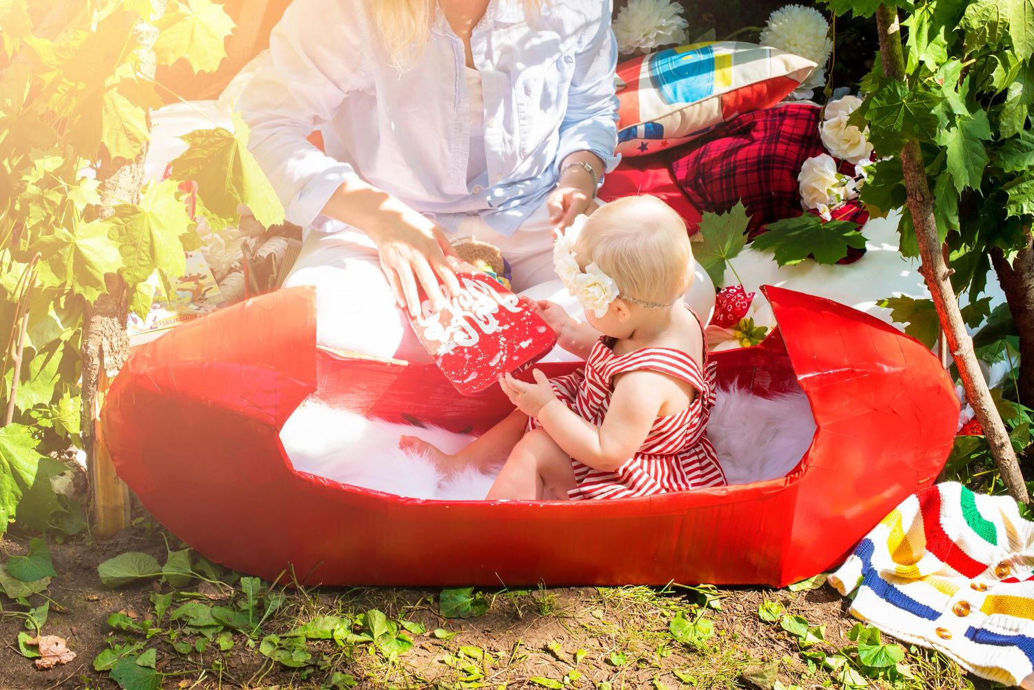 Baby girl in red striped dress sitting in red canoe prop outdoors in sunny garden setting.