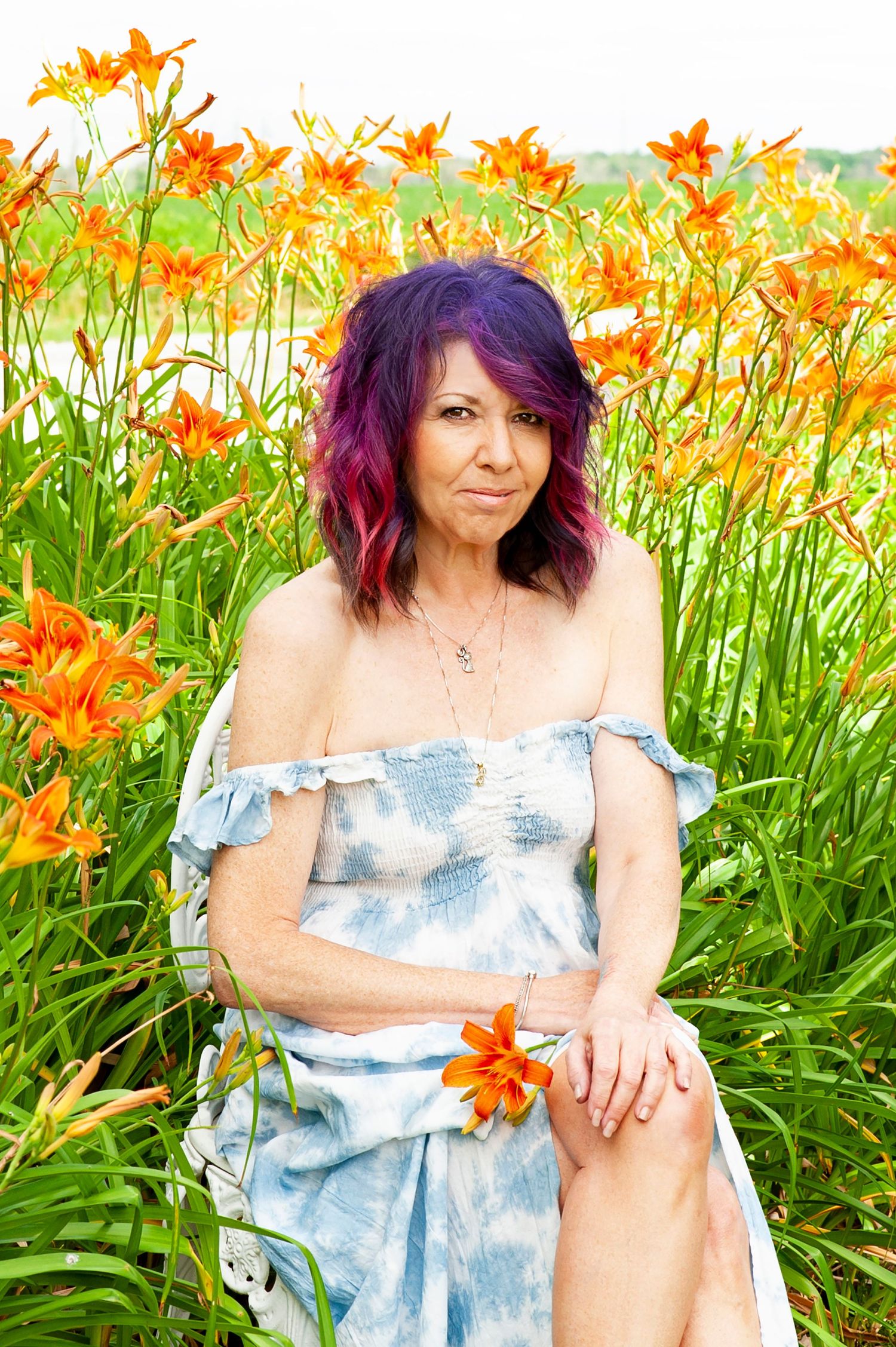 Woman with purple hair sitting among vibrant orange day lilies in field.