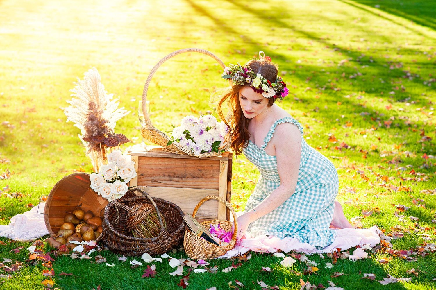 Young woman in floral crown and gingham dress arranging flowers in wooden cart on grass.