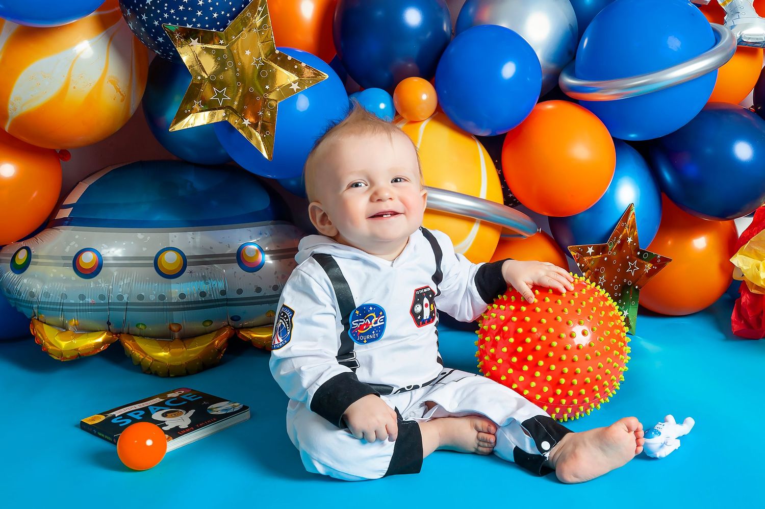 Baby in astronaut costume sits surrounded by space-themed balloons and planet decorations at a first birthday party.