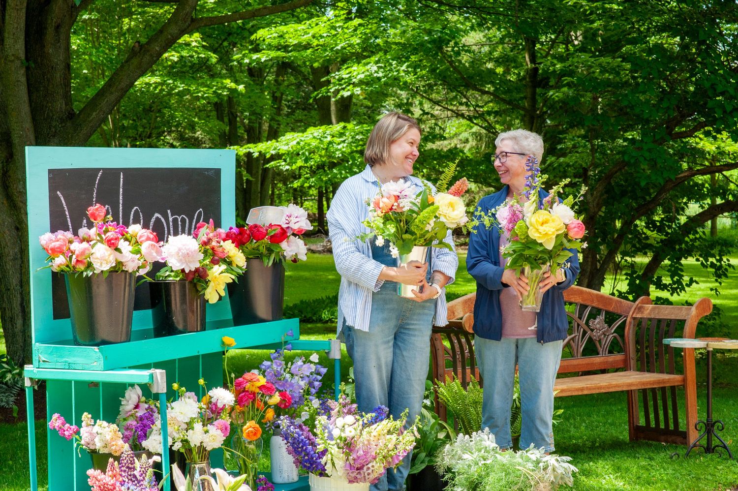 Two women arranging colorful fresh flowers at outdoor market stand with welcome chalkboard sign.