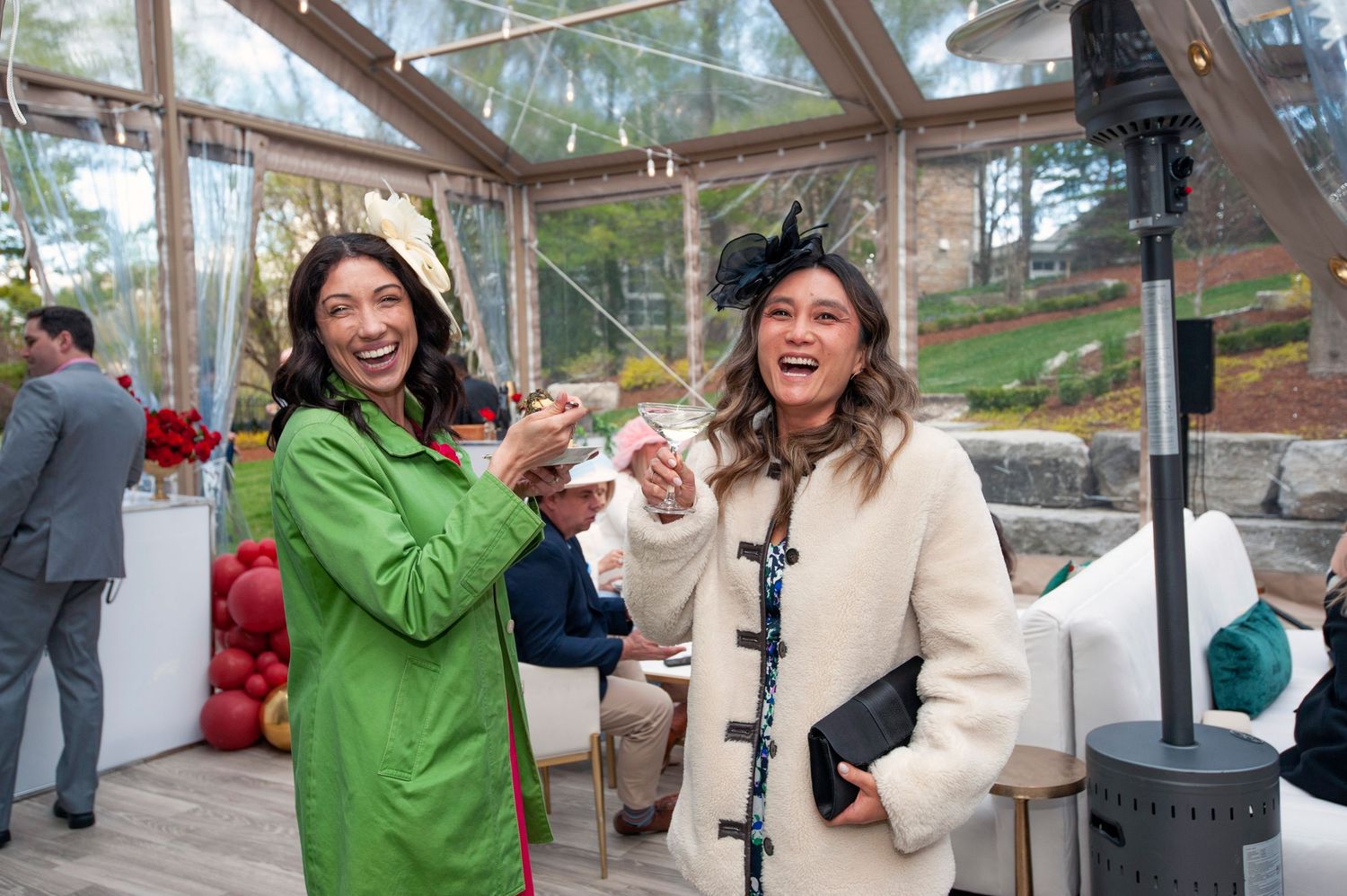 Two women laughing and toasting drinks inside a glass pavilion at a festive Kentucky Derby party event.