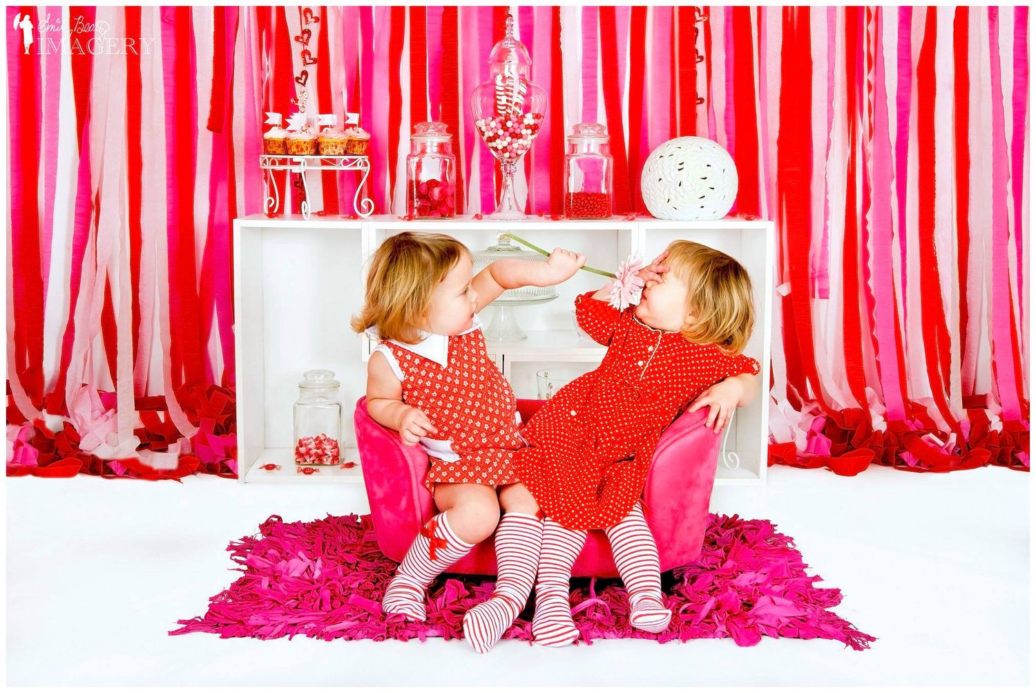 Two toddler girls in red dresses sit together on a pink chair against a festive red and pink streamer backdrop.