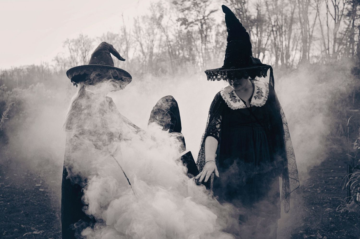 Black and white photo of two witches in hats surrounded by dramatic smoke in an outdoor setting.