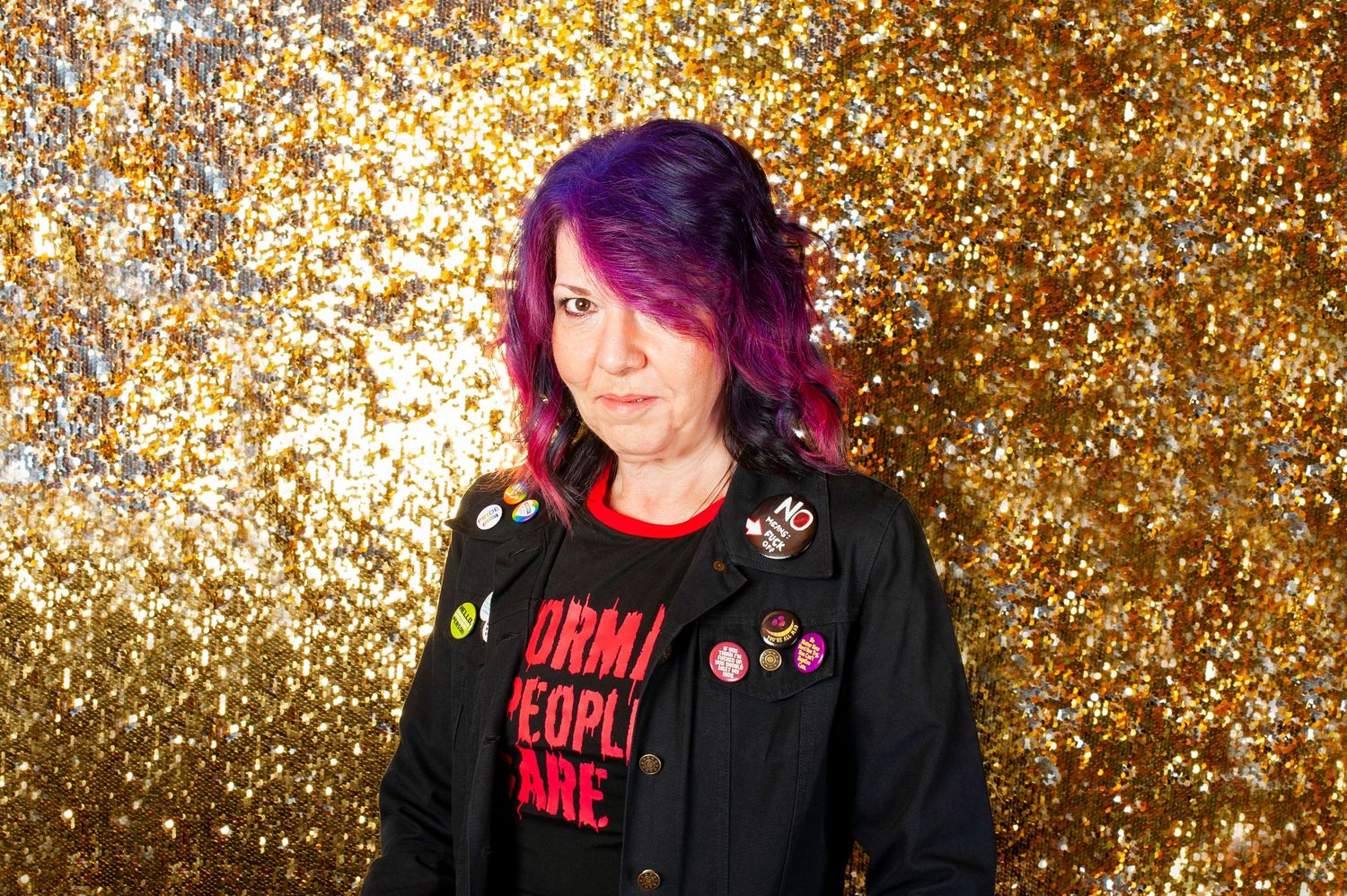 Woman with purple hair wearing black jacket against gold glitter background.