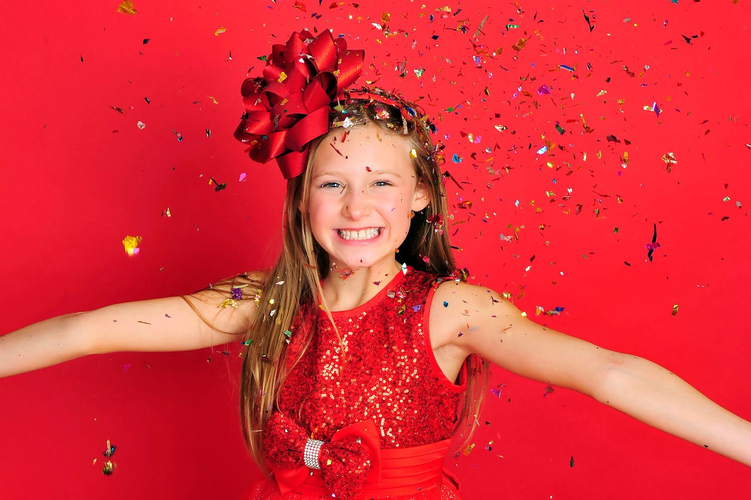 Happy girl in red sequin dress with bow headband, arms spread wide with colorful confetti falling.
