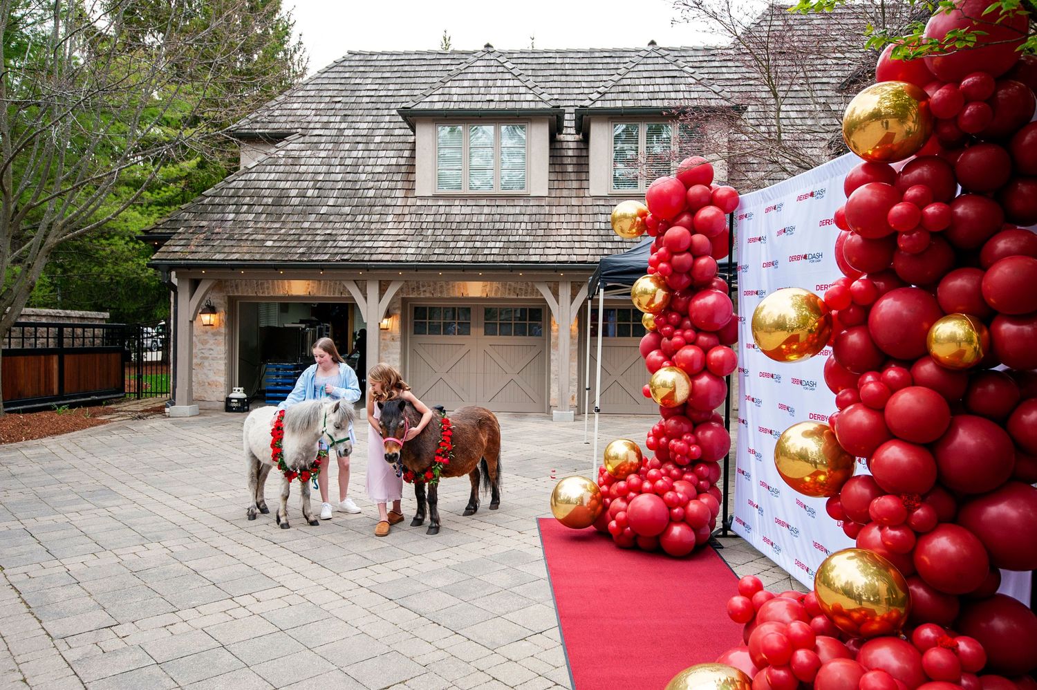 Girl riding a pony on a red carpet beside balloon arch decorations at an elegant outdoor party venue.