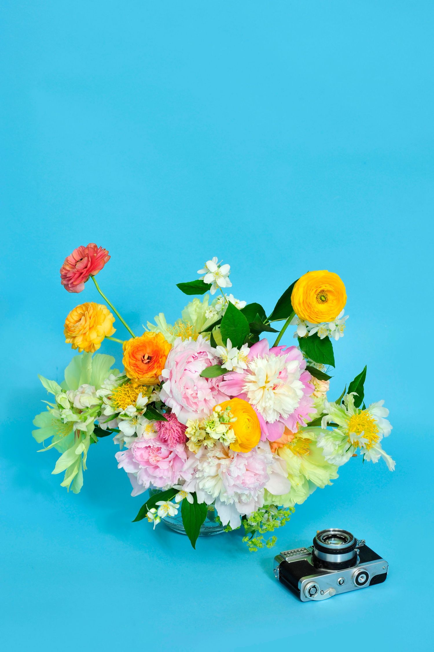Colorful spring flower bouquet with peonies and ranunculus in glass vase beside vintage camera on blue background.