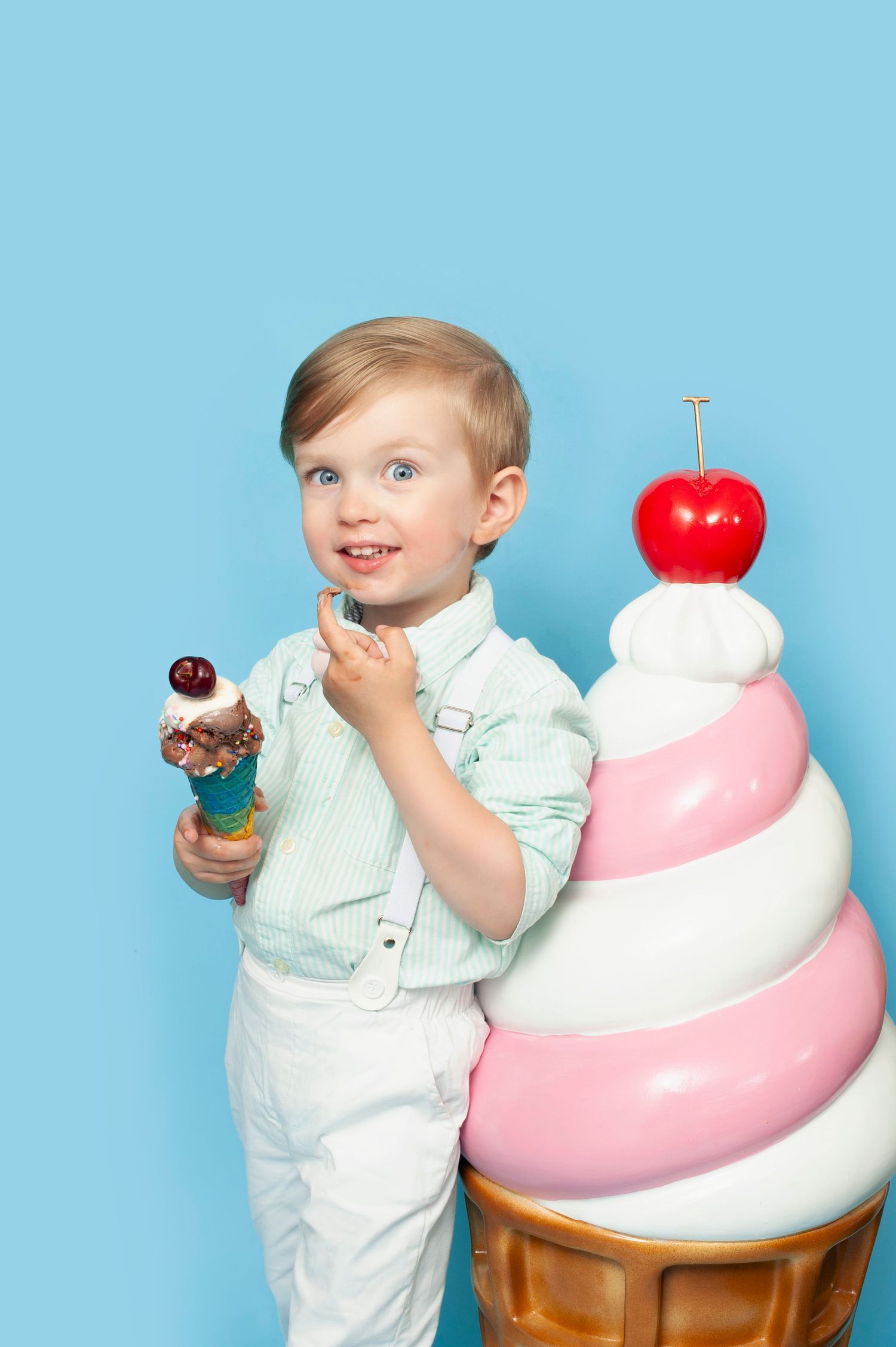 Smiling blonde toddler boy holds an ice cream cone beside a giant decorative ice cream on blue background.