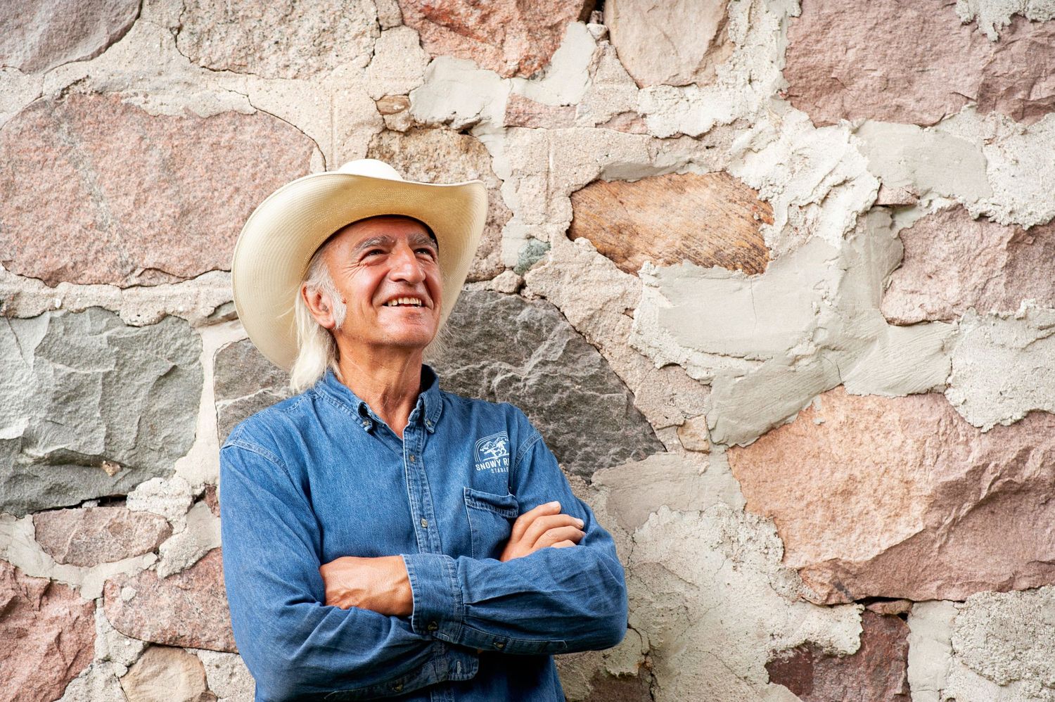 Smiling older man in cowboy hat and denim shirt leans against rustic stone wall with arms crossed.