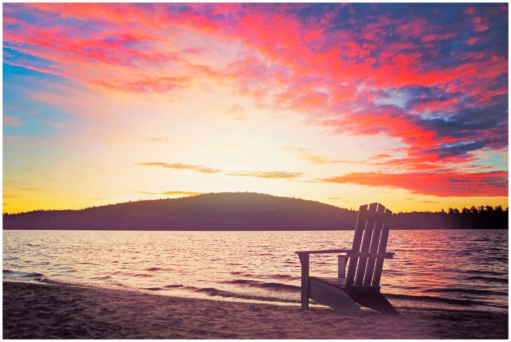 Adirondack chair on sandy beach at vibrant sunset with pink and purple sky reflecting over calm lake and distant hills.