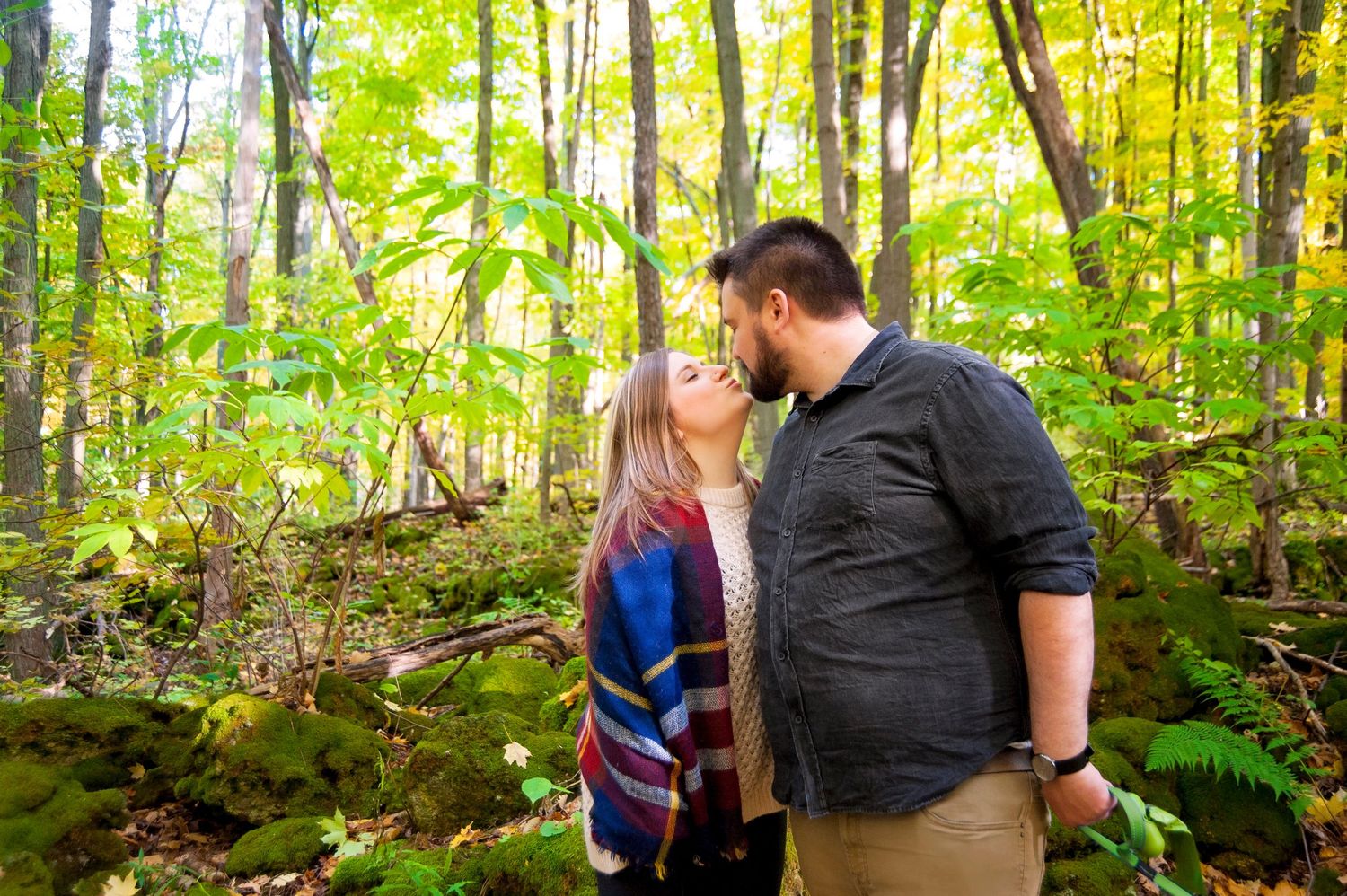 Couple sharing a tender kiss surrounded by lush green trees in a peaceful woodland setting.