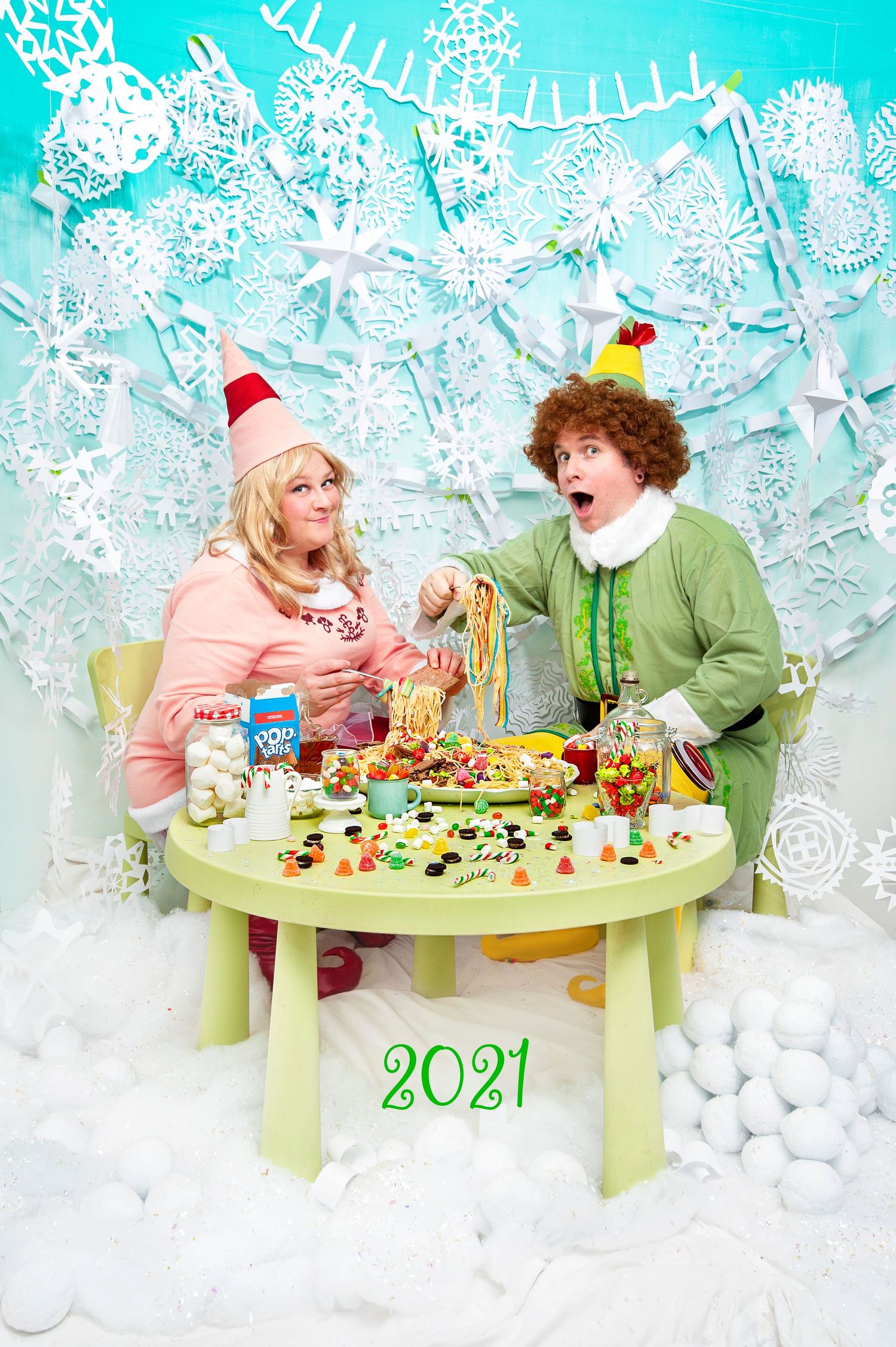 Two people dressed as Christmas elves sit at a candy-covered table in a festive winter wonderland set marked 2021.