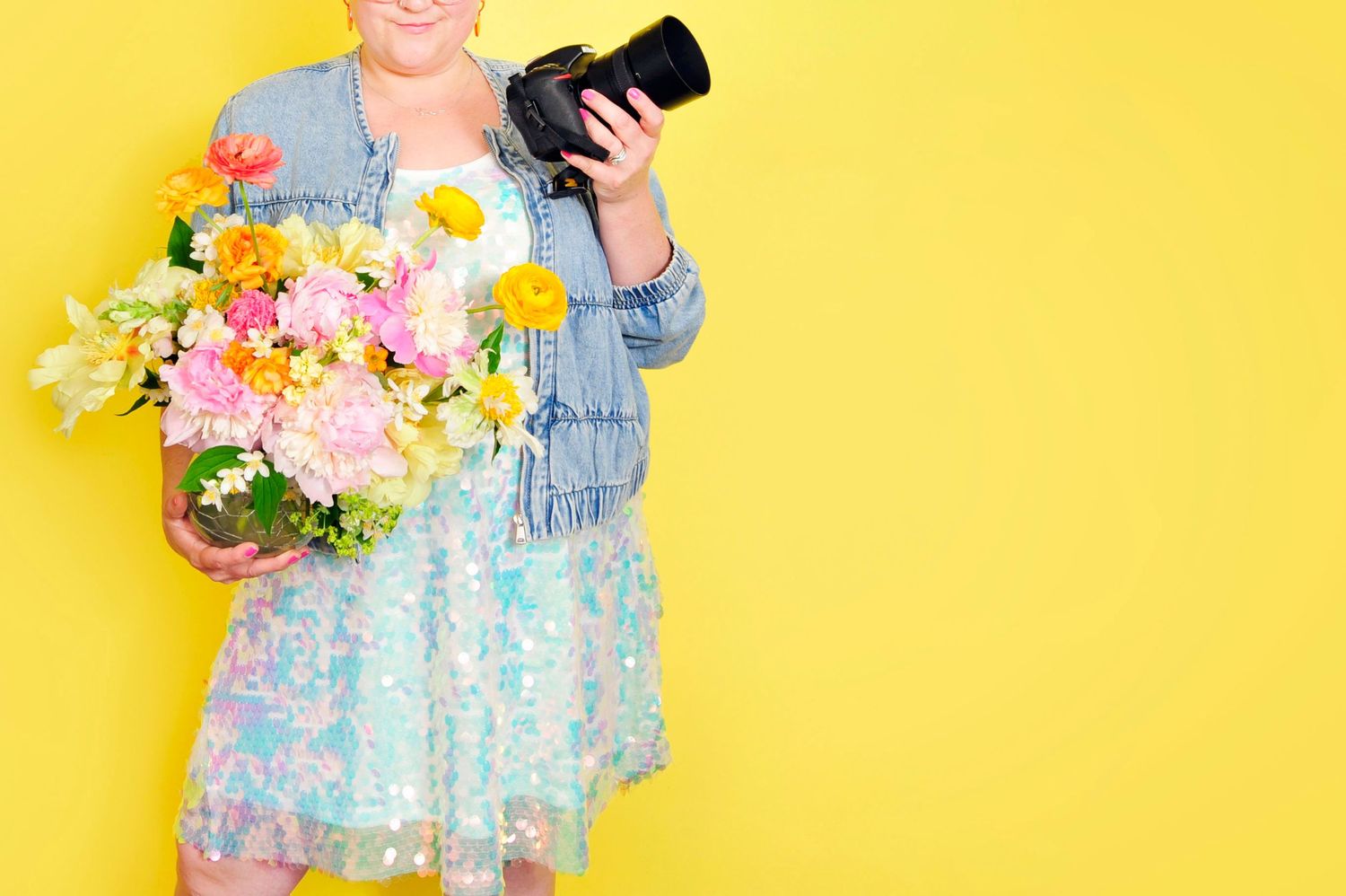 Woman in sequin dress holding colorful flower bouquet and camera against bright yellow background.