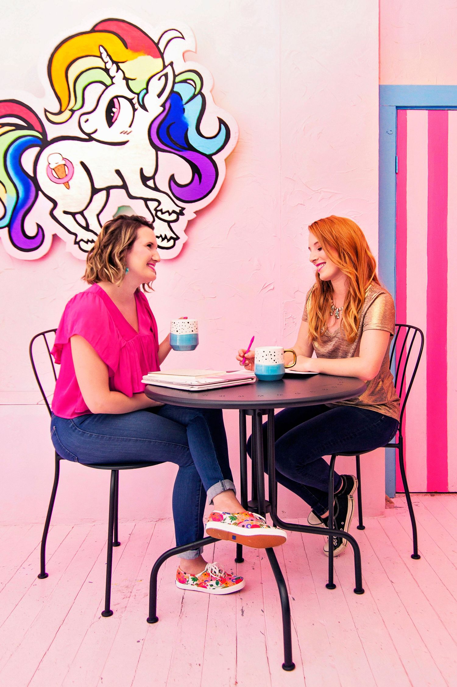 Two women laughing and chatting over coffee at a table with a rainbow unicorn mural on a pink wall.
