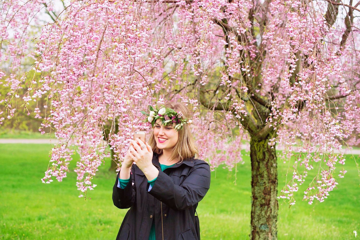 Woman smiling under a blooming pink cherry blossom tree wearing a floral crown in spring.